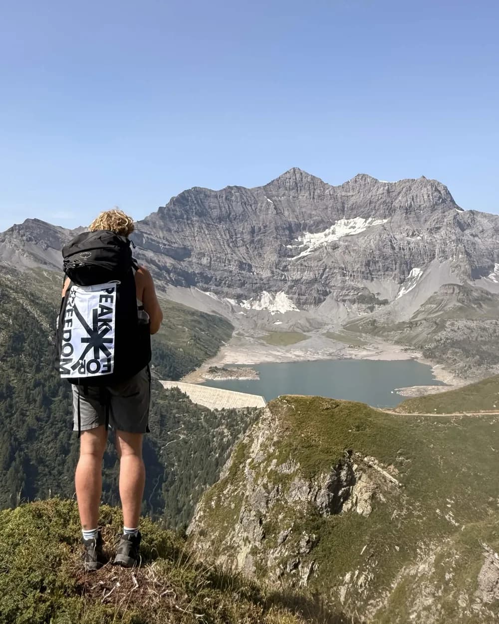 Hiker seen from behind with the Peak For Freedom flag on a backpack, above a turquoise mountain lake
